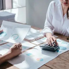 Dwie osoby analizują wykresy i raporty, jedna korzysta z kalkulatora. Rozważają amortyzację jednorazową, planując finanse.