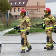 Strażacy w akcji. Czy wiesz, ile zarabia strażak? Jeden z nich kieruje strumień wody na pojazd, drugi niesie wąż.