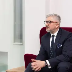 Mężczyzna w garniturze i okularach siedzi na czerwonym fotelu, patrząc w bok.