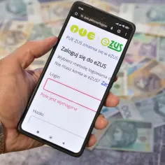 Dłoń trzyma smartfon z ekranem logowania do eZUS, na tle rozłożonych polskich banknotów.