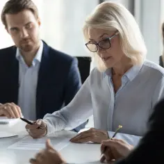 Kobieta w okularach omawia dokumenty, ucząc się, jak zarządzać zespołem pracowników. Obok siedzą mężczyźni w garniturach.