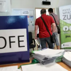 Niebieska teczka z napisem OFE stoi na biurku obok stosów dokumentów. W tle mężczyźni wchodzą do urzędu ZUS, gdzie wisi baner zachęcający do korzystania z platformy PUE.