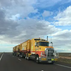 Żółta ciężarówka typu road train z wieloma przyczepami jedzie pustą drogą pod chmurami.