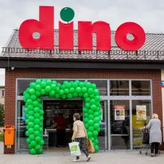 Wejście do polskiego sklepu Dino ozdobione zielonymi balonami. Klienci wchodzą i wychodzą, robiąc zakupy.