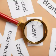 Lupa powiększa słowo "Law" wśród kart z napisami: "Trademark", "Trade Secret", "Copyright", "Intellectual Property", "Brand", "License". To symbol analizy prawnej w dziedzinie **wnip**.