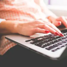 Dłonie z czerwonymi paznokciami piszą na laptopie. To praca copywritera, tworzącego angażujące teksty.