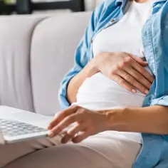 Kobieta w ciąży na kanapie z laptopem, przygotowująca się do urlopu macierzyńskiego i rodzicielskiego.