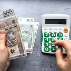 Dłoń trzyma banknoty 200 zł i 100 zł, obok kalkulator z wyświetloną kwotą 50000. Obrazek symbolizuje dyskusję o wynagrodzeniu minimalnym.