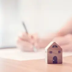 Mały drewniany domek na stole, obok podpisująca umowa kredytowa.