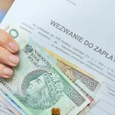 Dłoń trzyma banknoty 100 i 200 zł na dokumencie "Wezwanie do zapłaty". Zastanawiasz się, ile zarabia komornik?