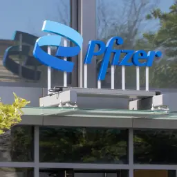 Niebieskie logo Pfizer na budynku. W tle odbijają się drzewa i niebo.