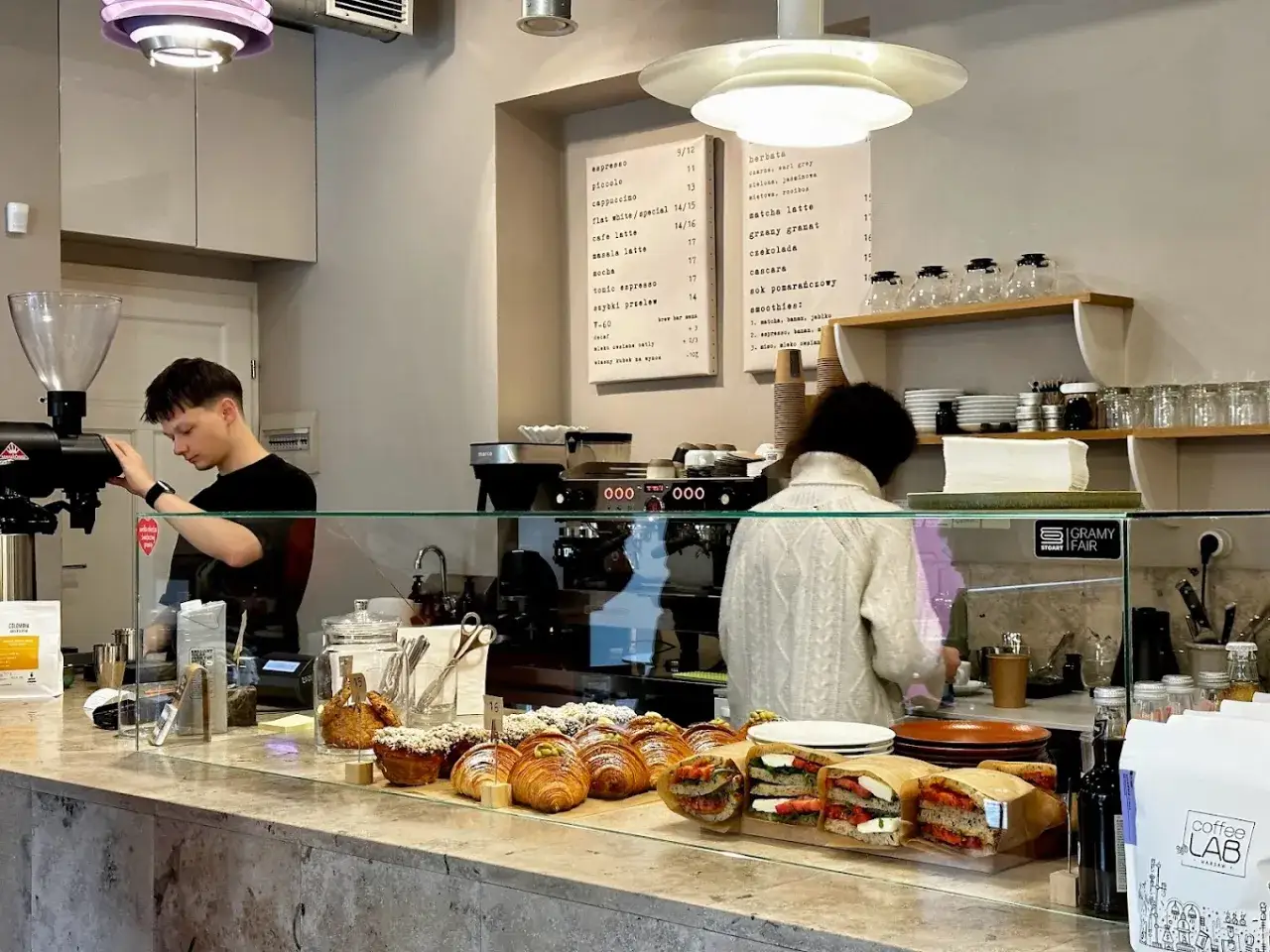 Młody barista przygotowuje kawę w kawiarni. Na ladzie świeże croissanty i kanapki.