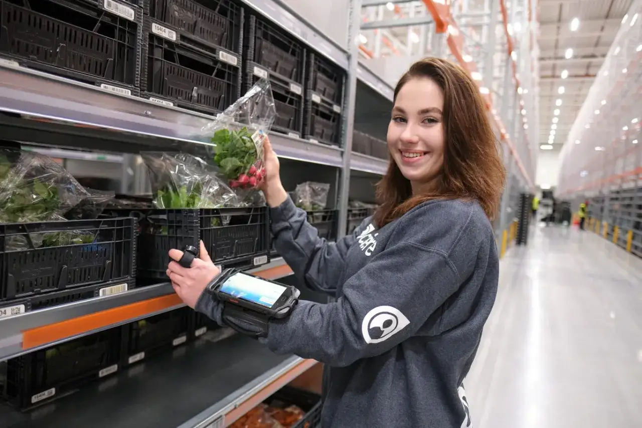 Uśmiechnięta pracownica kompletuje zamówienie w magazynie. To jej praca w Holandii, gdzie skanuje produkty i przygotowuje je do wysyłki.