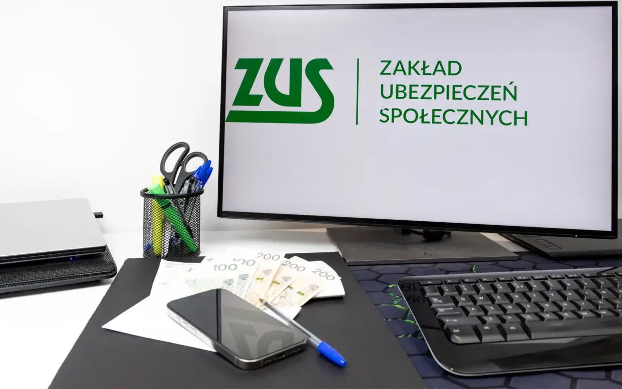 Biurko z logo ZUS na monitorze, telefonem, pieniędzmi i artykułami piśmienniczymi. Idealne miejsce na rozpoczęcie stażu.