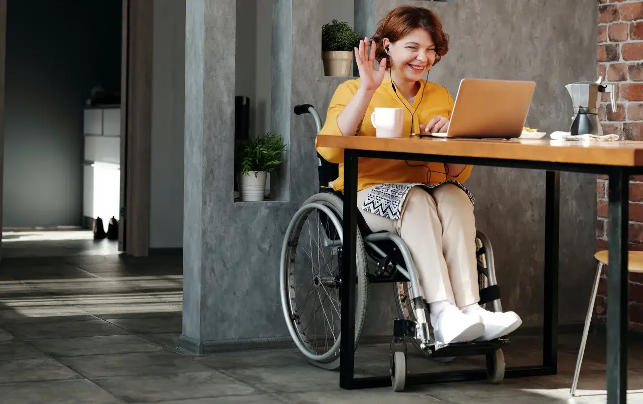 Kobieta na wózku inwalidzkim wita się przez laptopa, pracując zdalnie. To jej dodatkowa praca, którą wykonuje z uśmiechem.