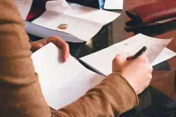 Osoba notuje na kartce, obok leżą dokumenty i monety. Może to być przygotowanie do złożenia wniosku o gwarancję bankową.