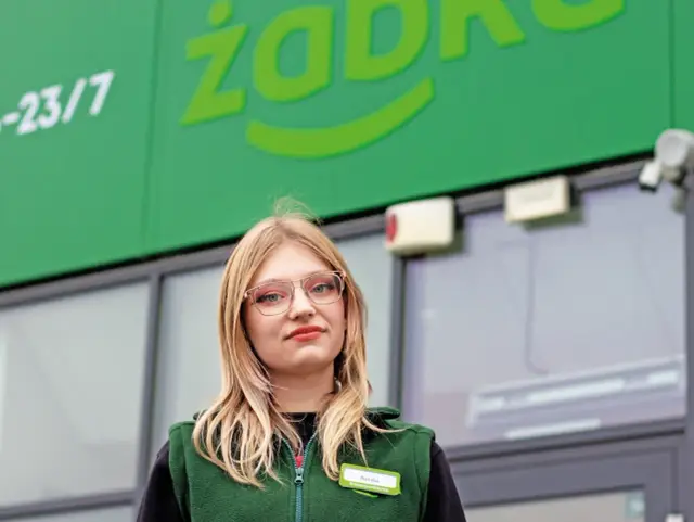 Natalia, pracownica Żabki, z uśmiechem pozuje przed sklepem.